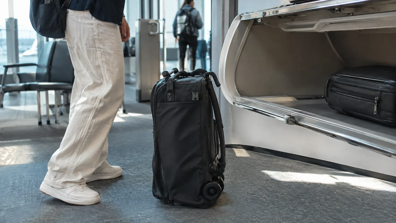 A compact travel stroller folded into a small carry-on bag next to an overhead bin