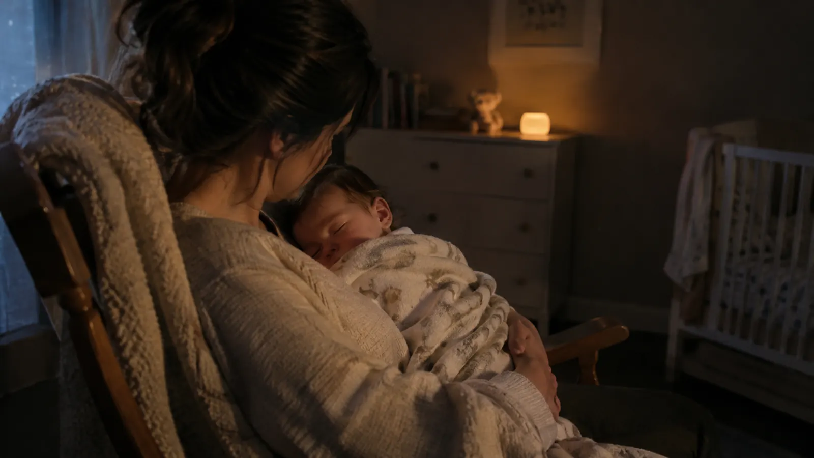 A parent holding a sleepy baby in a rocking chair in a dim nursery