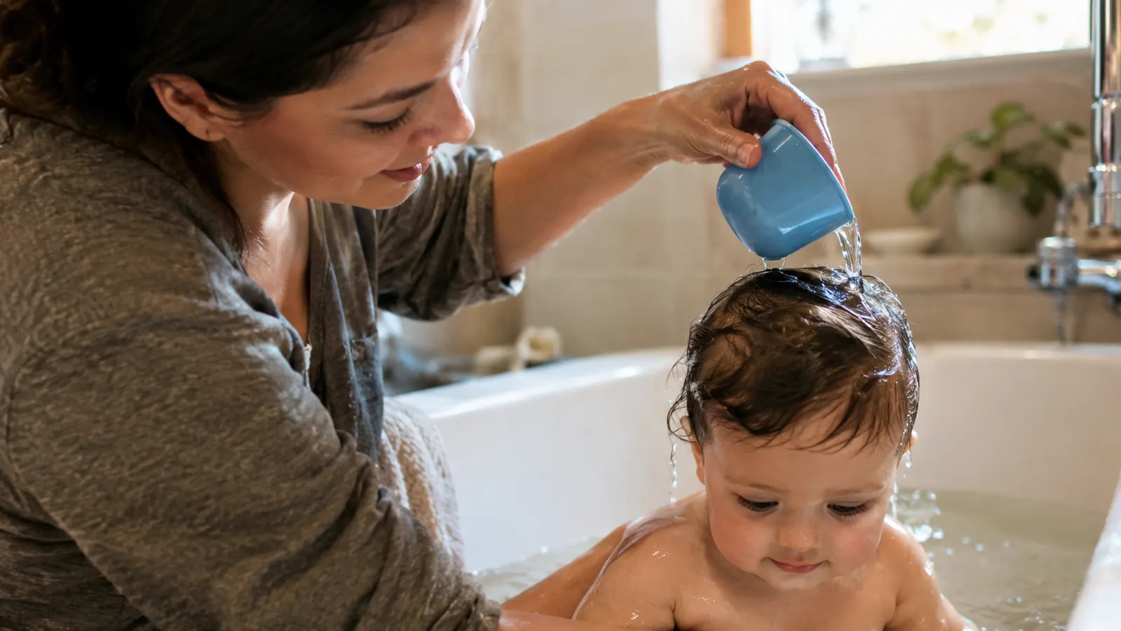 A parent rinsing a baby's hair with a small cup in a bathtub